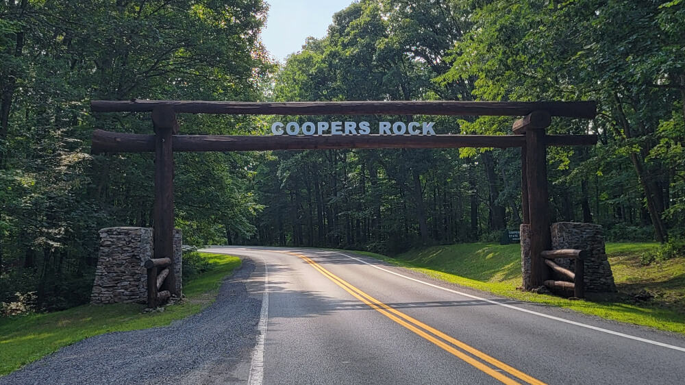 A sign made out of wood spans a two lane road and reads "Coopers Rock"