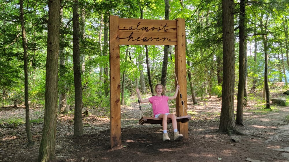 A young girl sits on a wooden swing. The sign above the swing reads "almost heaven"