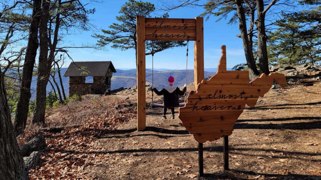 Young girl sits on a swing with "almost heaven" written at the top of the swing. There is a wooden cutout in the shape of West Virginia with "almost heaven" written on it in the foreground