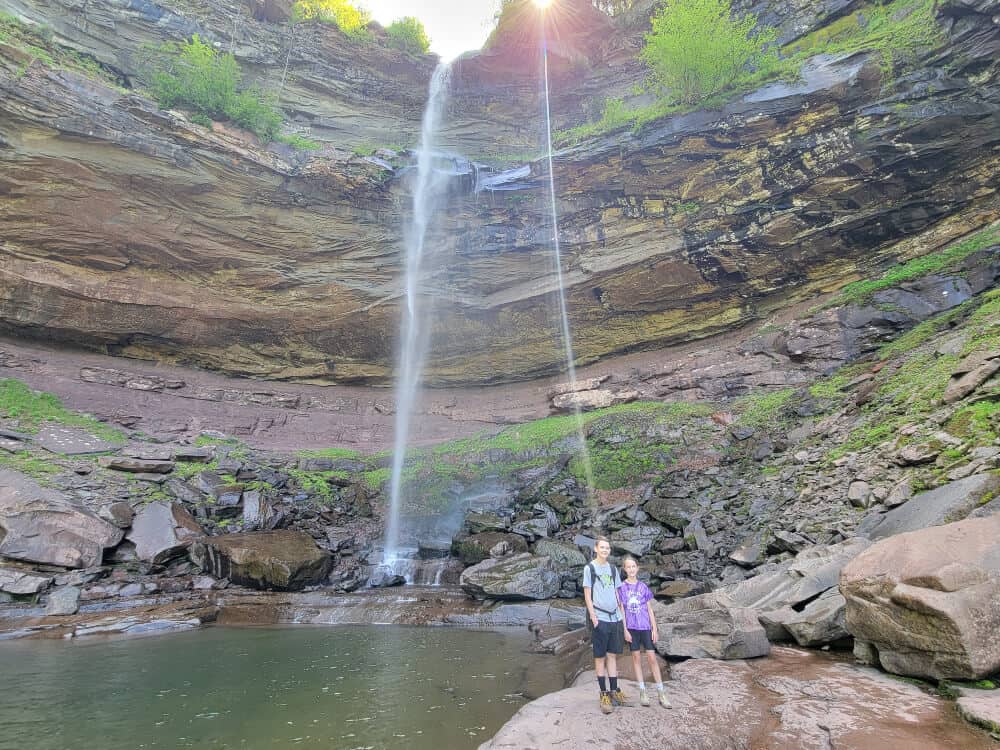 two kids stand in front of a thin waterfall with rock walls surrounding