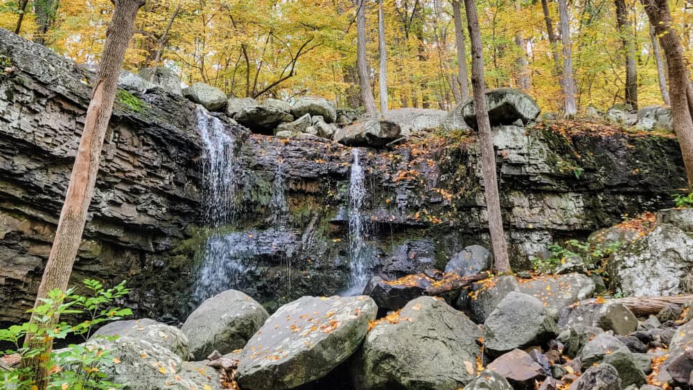 A waterfall with large boulders at the base and trees with fall leaves in the background