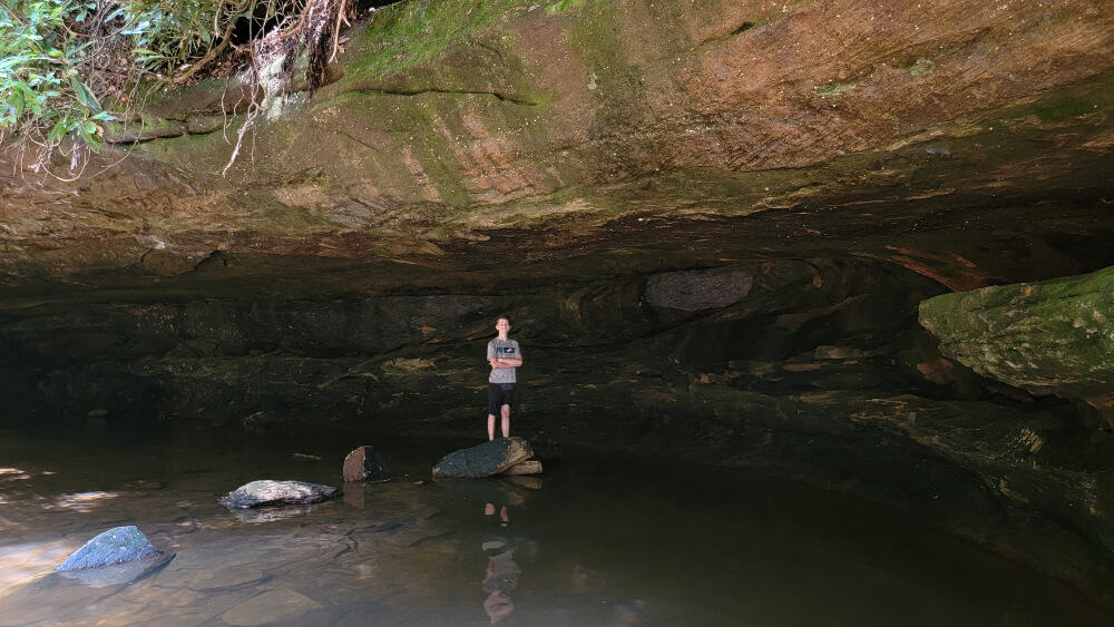 A boy stands underneath a rock that forms a "grotto"  in shallow water