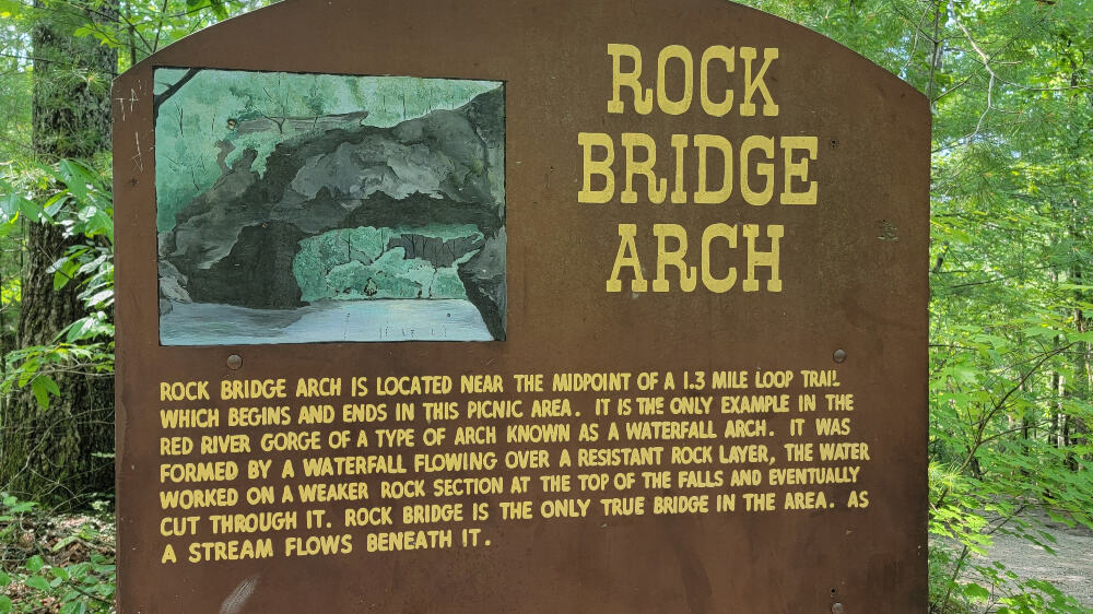 sign says "rock bridge arch is located near the midpoint of a 1.3 mile lop trail which begins and ends in this picnic area. it is the only example in the red river gorge of a type of arch known as a waterfall arch. it was formed by a waterfall flowing over a resistant rock layer, the water worked on a weaker rock section at the top of the falls and eventually cut through it. rock bridge is the only true bridge in the area as the a stream flows beneath it."