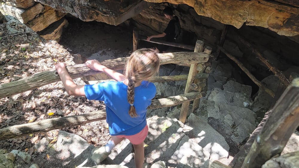 A young girl walks down a stone staircase using a wooden railing into a cave