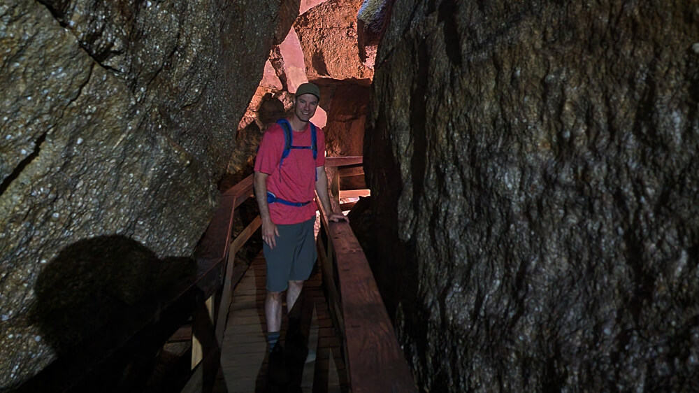 A man wearing a red shirt and gray shorts stands on a boardwalk path in a cave
