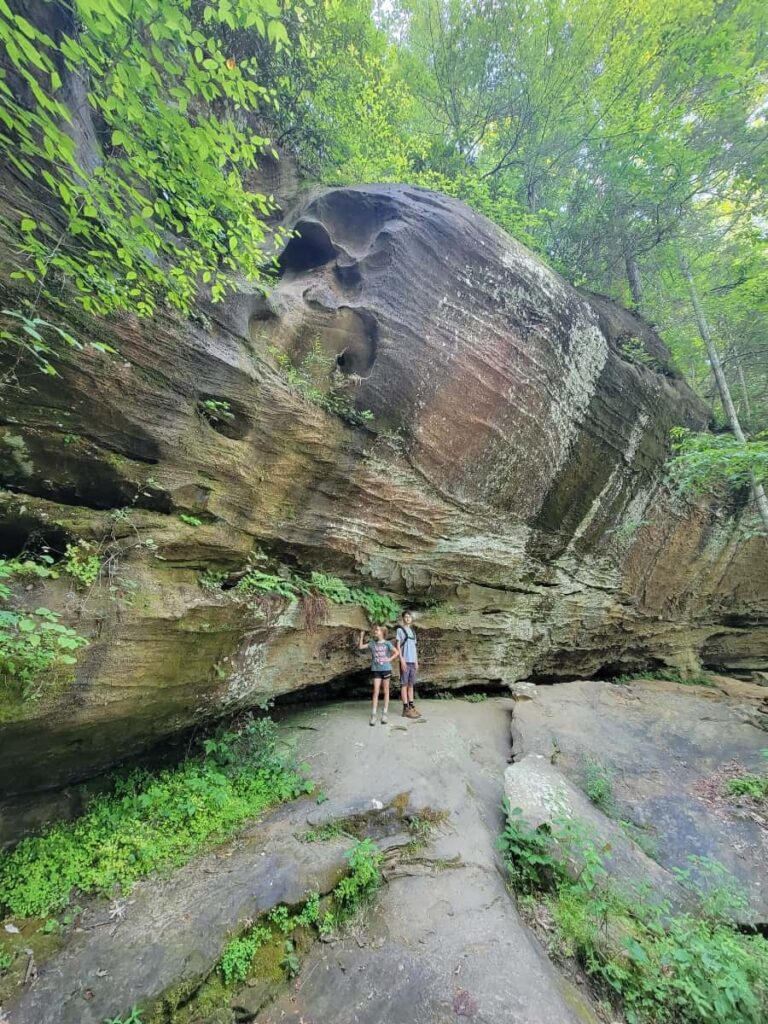 two kids stand in front of large stone wall at the base of sky bridge arch in red river gorge