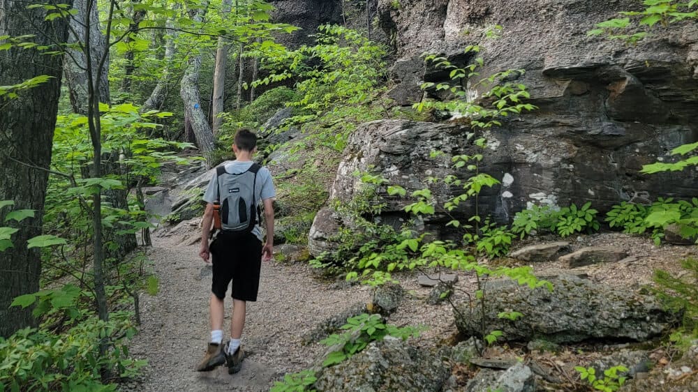 Boy walks on a hiking trail with trees to the left and large rocks to the right