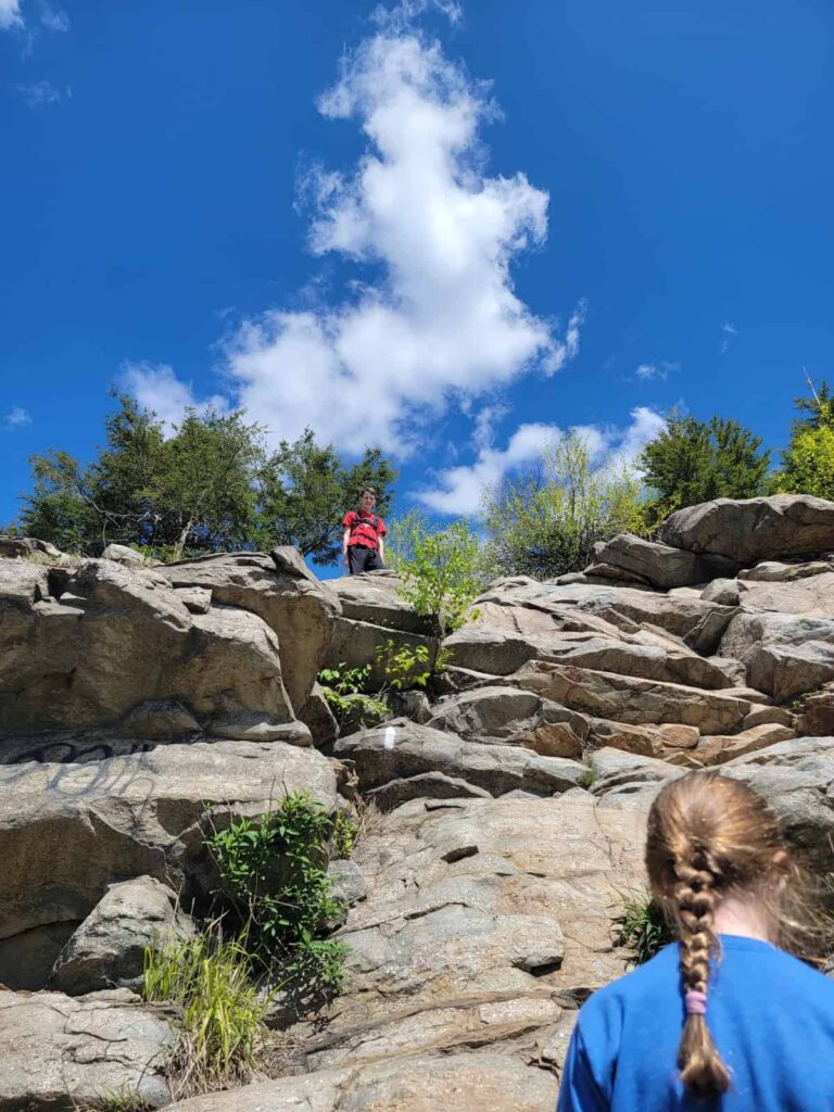 A girl stands below as a boy looks over a steep rock scramble