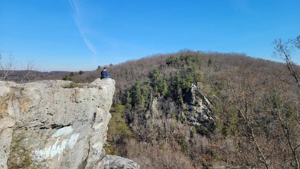 A man sits on the edge of a cliff with a small mountain in the distance at Rocks State Park in Maryland