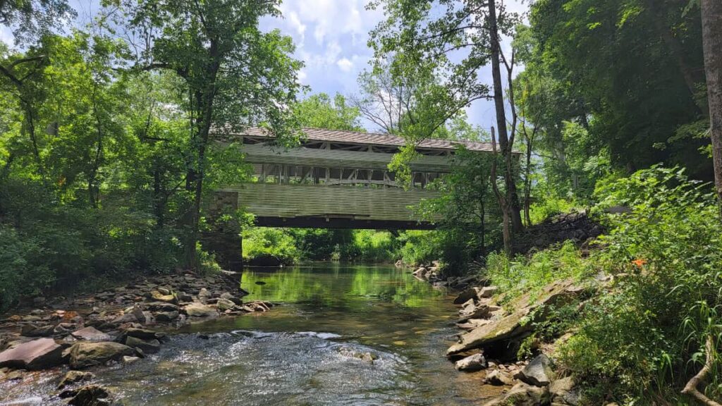 a tan covered bridge spans across a shallow creek