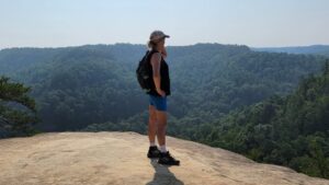 woman stands on rock looking out over a vista wearing blue shorts, a tank top, and hat