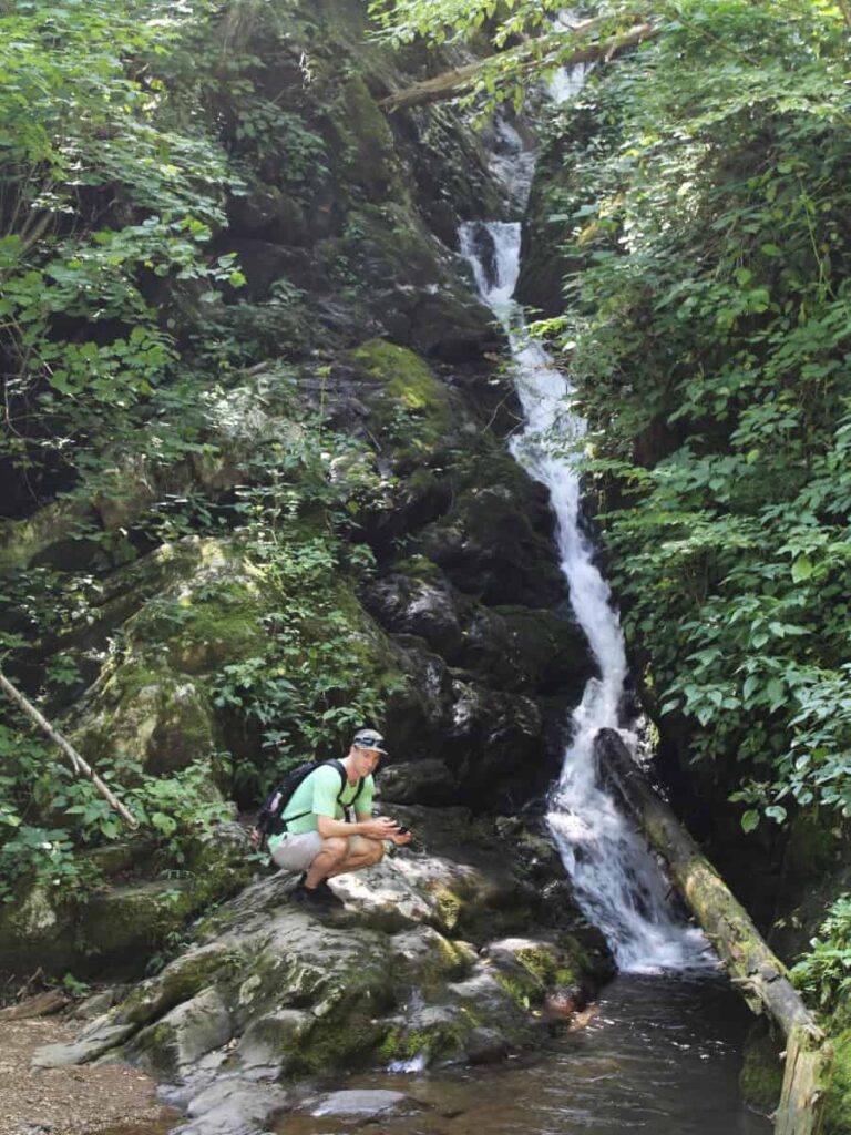 A man sits on a rock next to a tall skinny waterfall at Dark Hollow Falls 