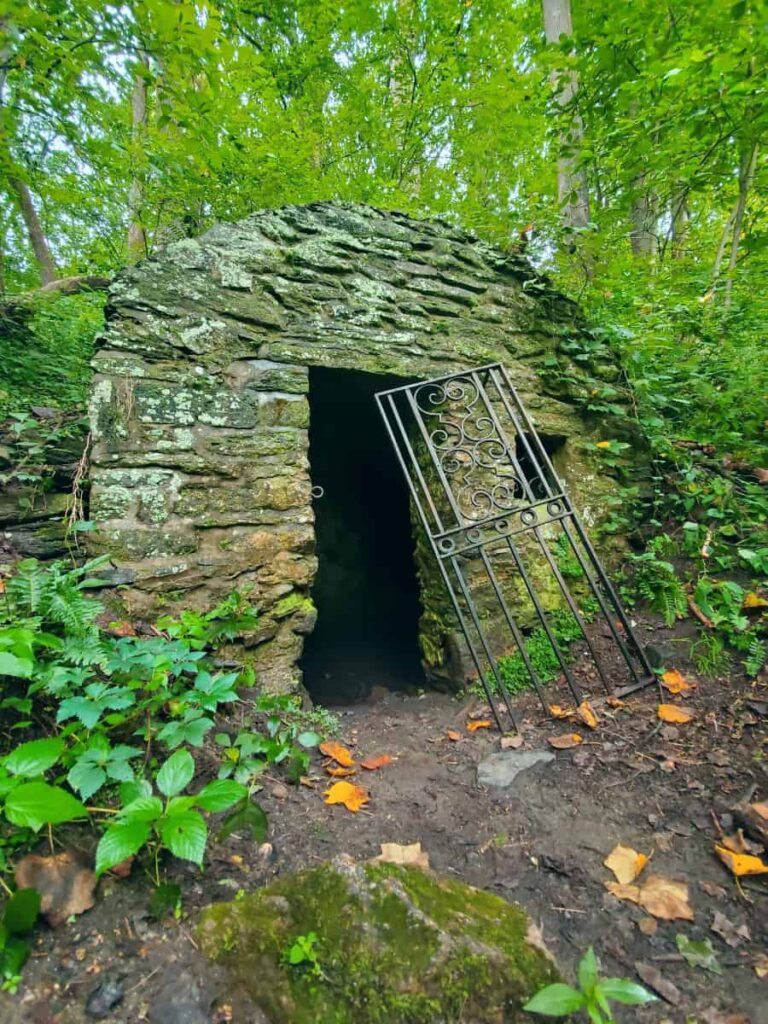 A stone spring house with a iron door propped up outside sits in the woods
