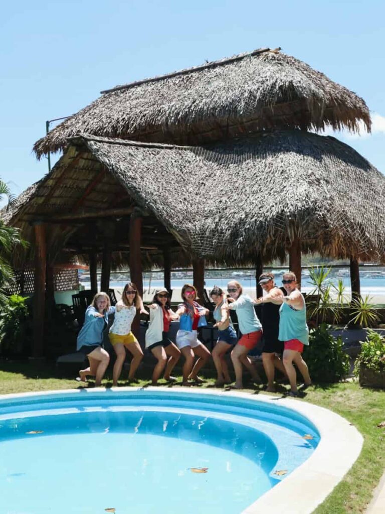 A group of women stand next to pool and a grass hut building