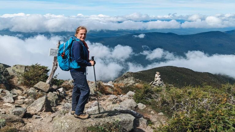 A woman stands on top of a mountain with clouds below her
