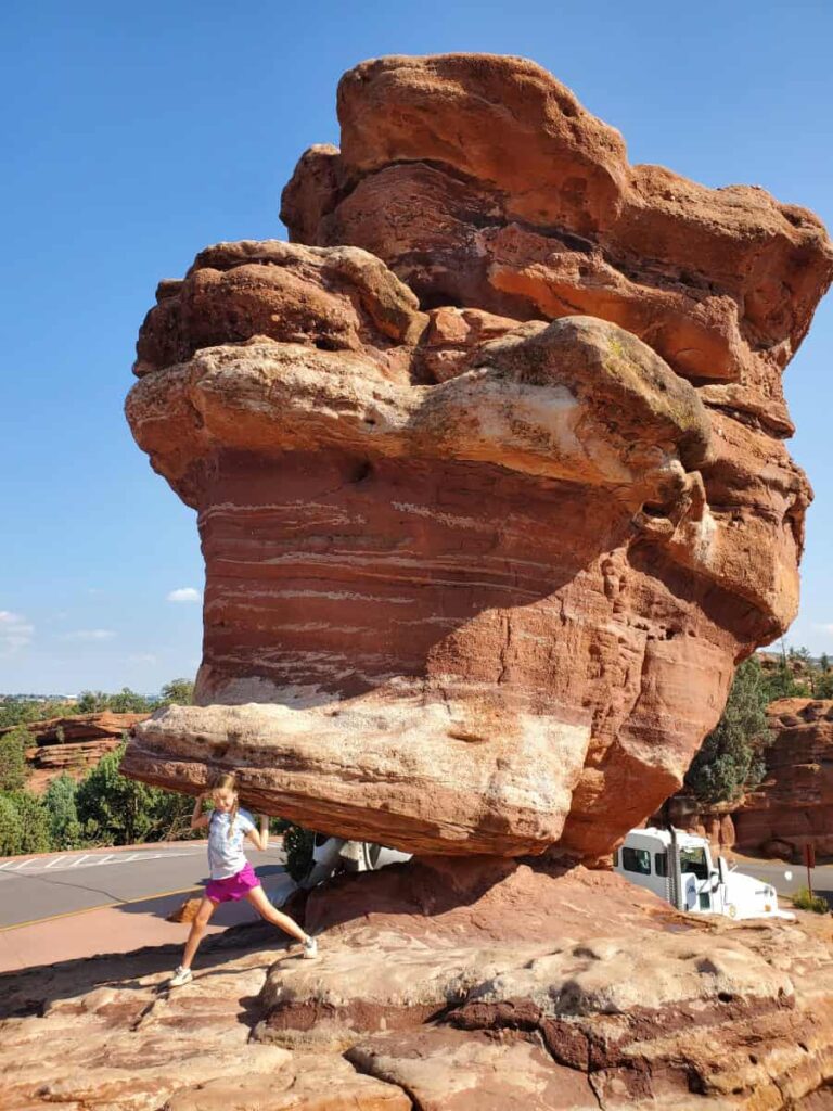 A young girls acts as though she is holding up a large rock formation (Balanced Rock)