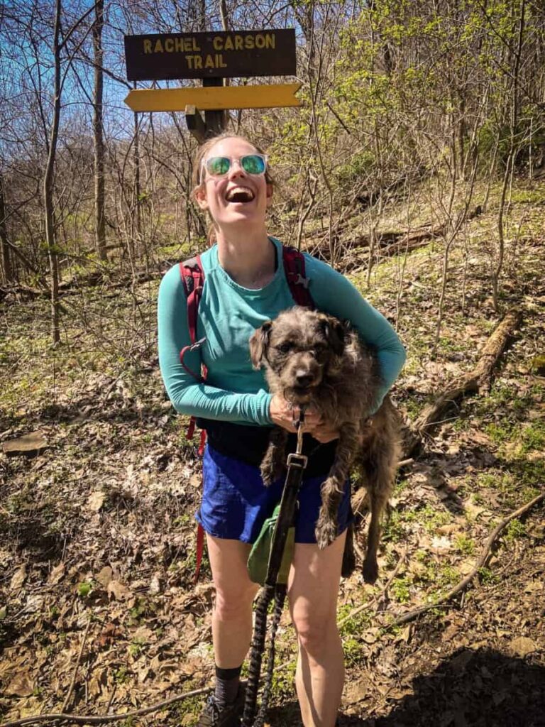 A woman holds up her dog while hiking on the Rachel Carson Trail
