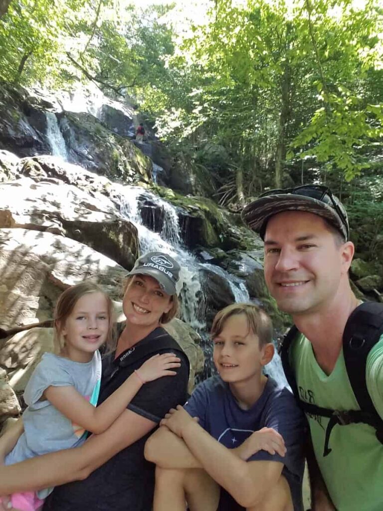 A family of four takes a selfie in front of the waterfall on the Dark Hollow Falls trail