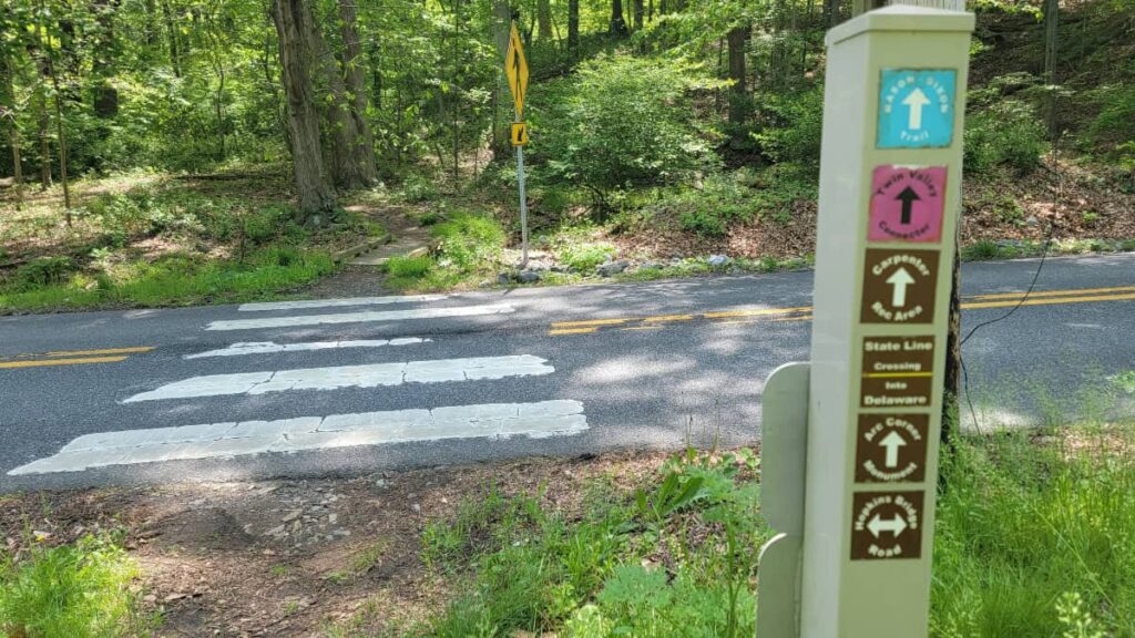 A crosswalk over a road in the distance with a trail sign indicating the state line between PA and DE