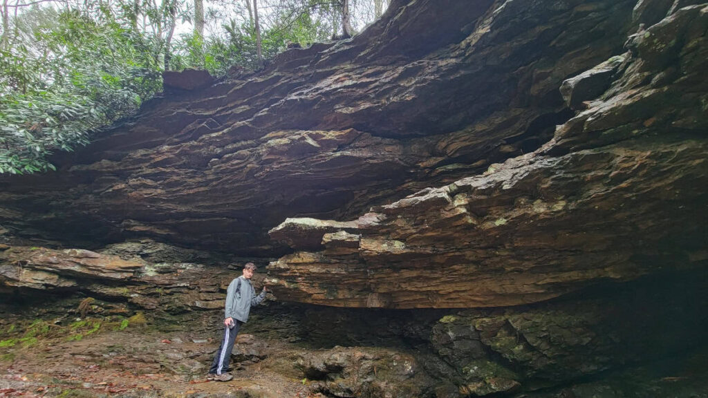 A young man stands below rock shelter at Trough Creek