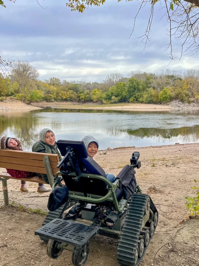 Two kids sit on a bench by a lake while a thirs child sits in a Track Chair wheel chair (made for off-roading)