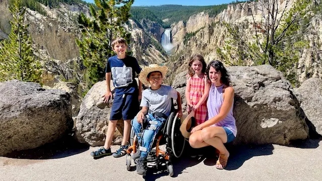 A mother crouches next to her 3 children (one of which is in a wheelchair) in front of a mountainous vista