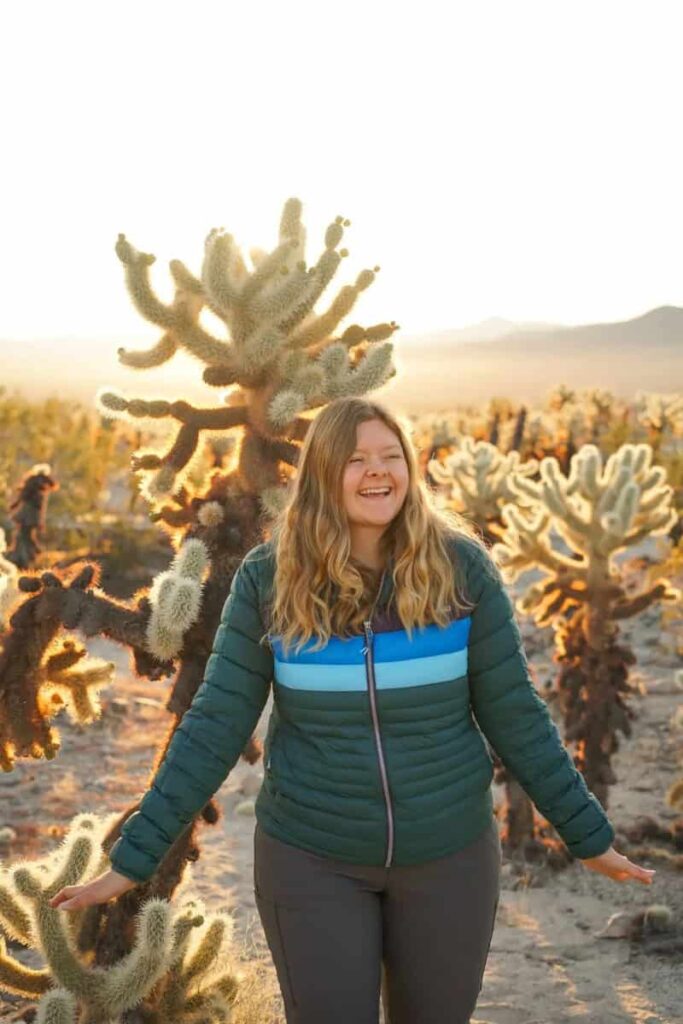 A woman wearing a green jackets stands in front of a Joshua Tree with the sun setting in the background