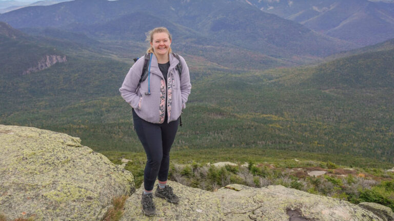 A woman stands on a rock with the mountains behind her in the distance
