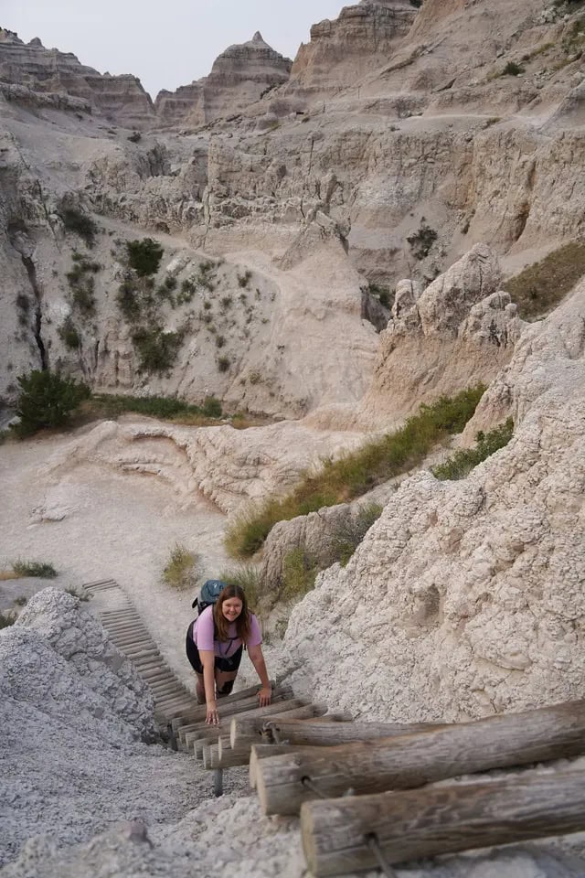 A woman climbs up a rope and wooden ladder along a steep rocky slope in the dessert