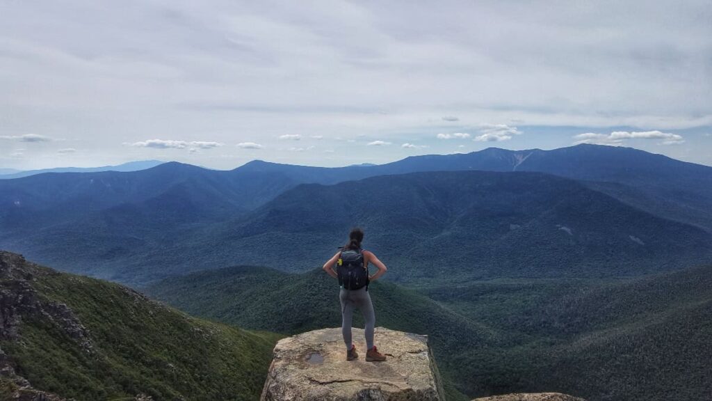 A woman stands on a large rock at the top of a mountain looking out to the distant landscape of trees