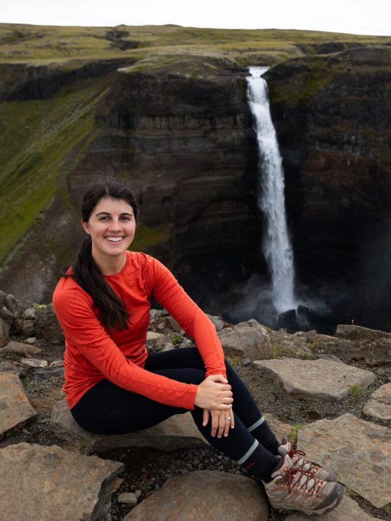 A woman sits on a rock in Iceland with a skinny waterfall behind her in the distance