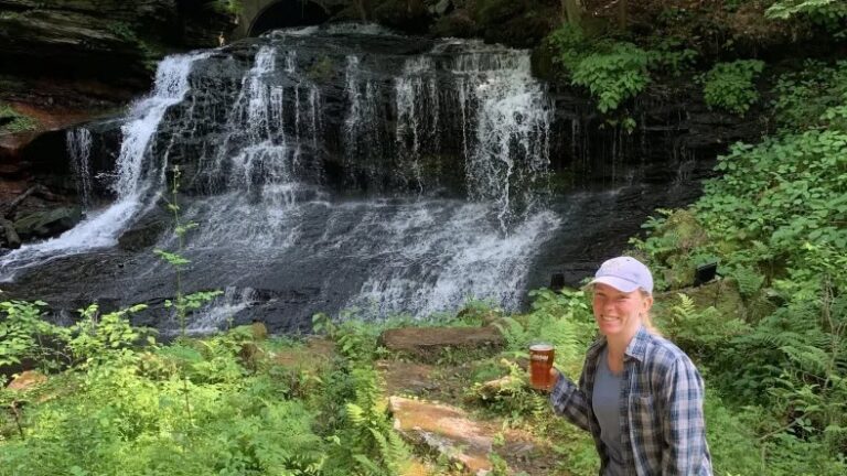 A woman stands in front of a waterfall with a glass of beer in her hand
