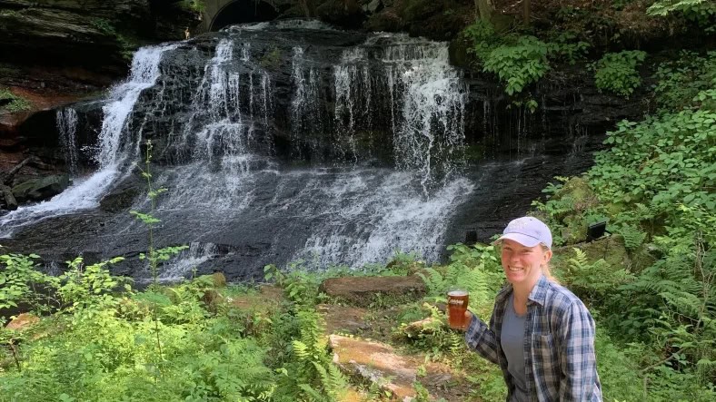 A woman stands in front of a waterfall with a glass of beer in her hand