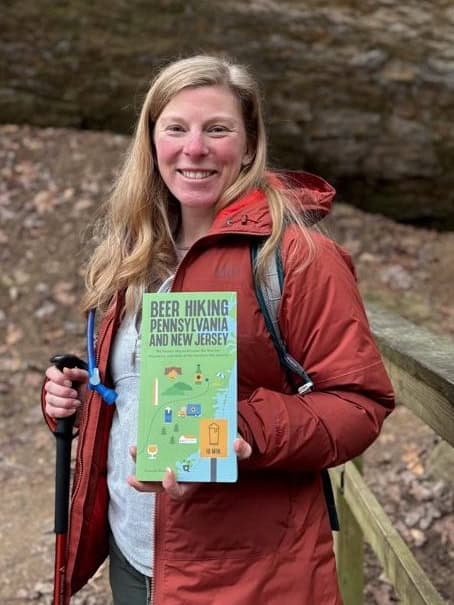 Hannah Powell stands on a hiking trail holding a copy of her book, Beer Hiking Pennsylvania and New Jersey