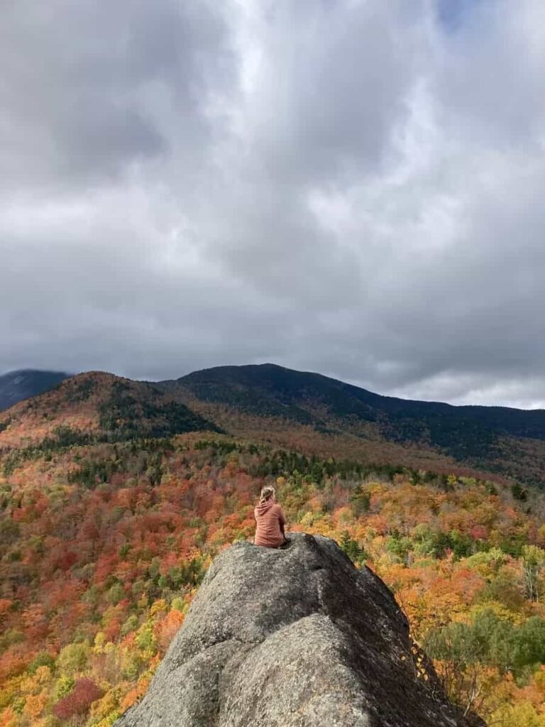 A woman sits on a rock outcropping overlooking fall colored trees