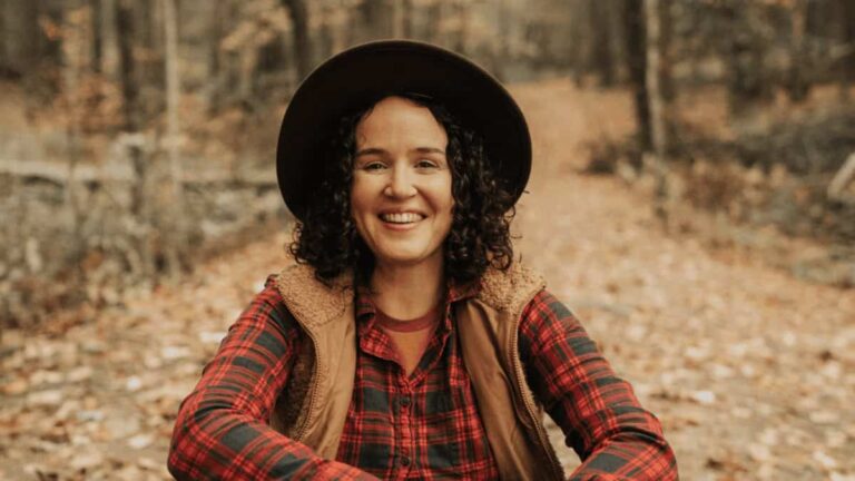 A woman sits on a leafy trail in the fall smiling and wearing a hat and vest
