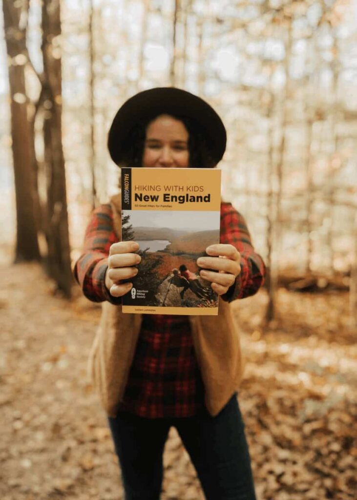 a woman stands on a hiking trail in the fall holding a trail guide up to the camera