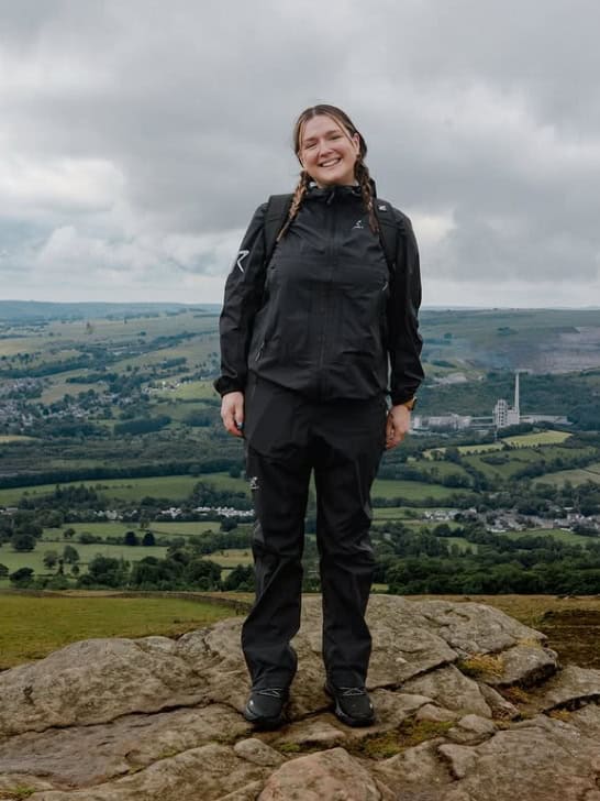 A woman stands at the top of a mountain smiling directly at the camera