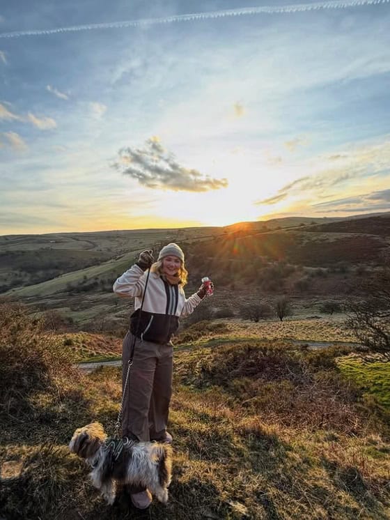 A woman stands at the top of a mountain with her dog watching the sunset