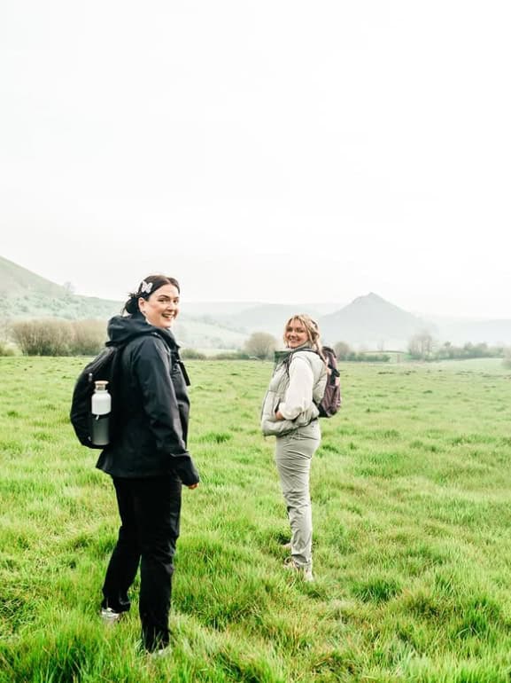 Two women stand in a grassy field with mountains in the distance