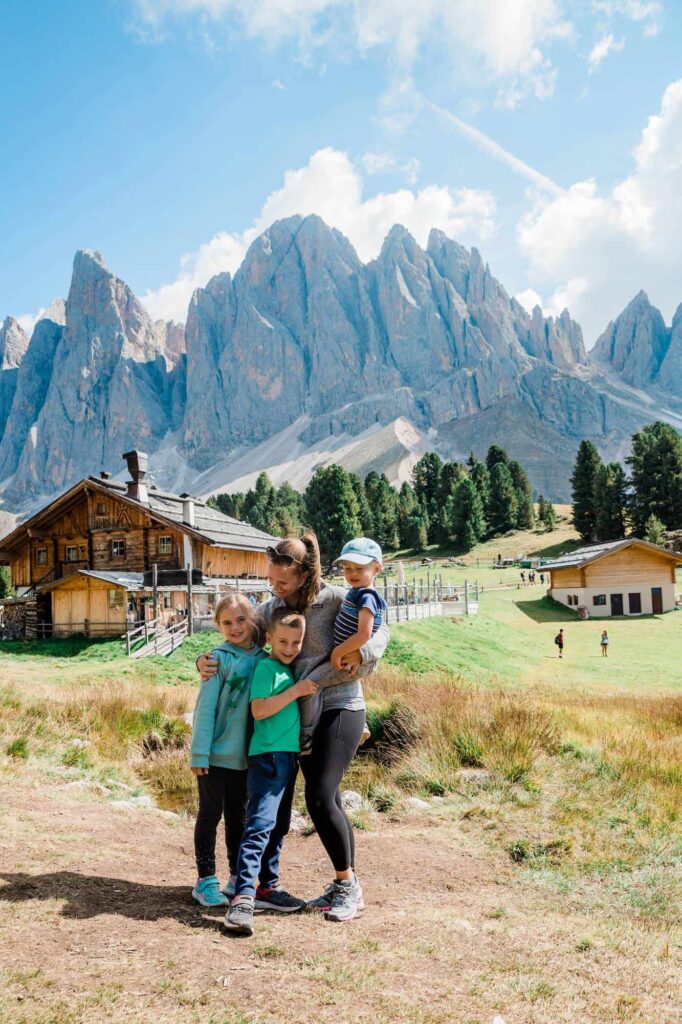 A woman hugs her three children standing front of a log cabin with mountains in the background