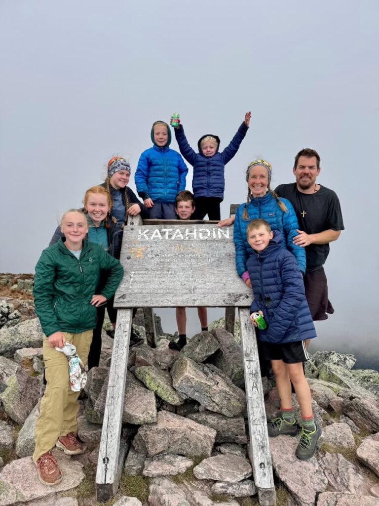 A family of seven kids and two adults surround the Katahdin sign on the Appalachian Trail