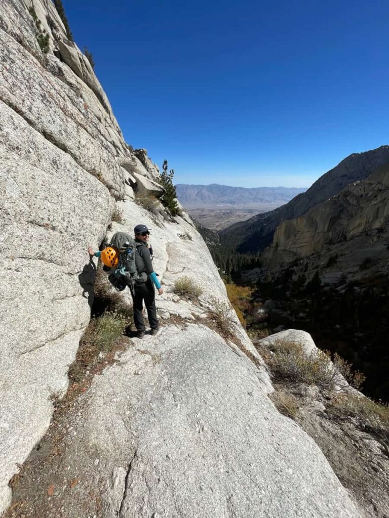 A woman stands on the side of a rocky cliff with her backpacking gear and helmet
