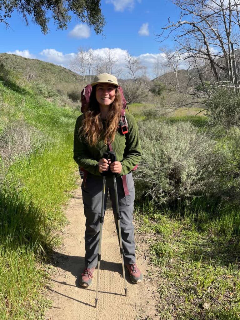 A woman stands with her hiking gear on a dirt trail with the mountains in the distance