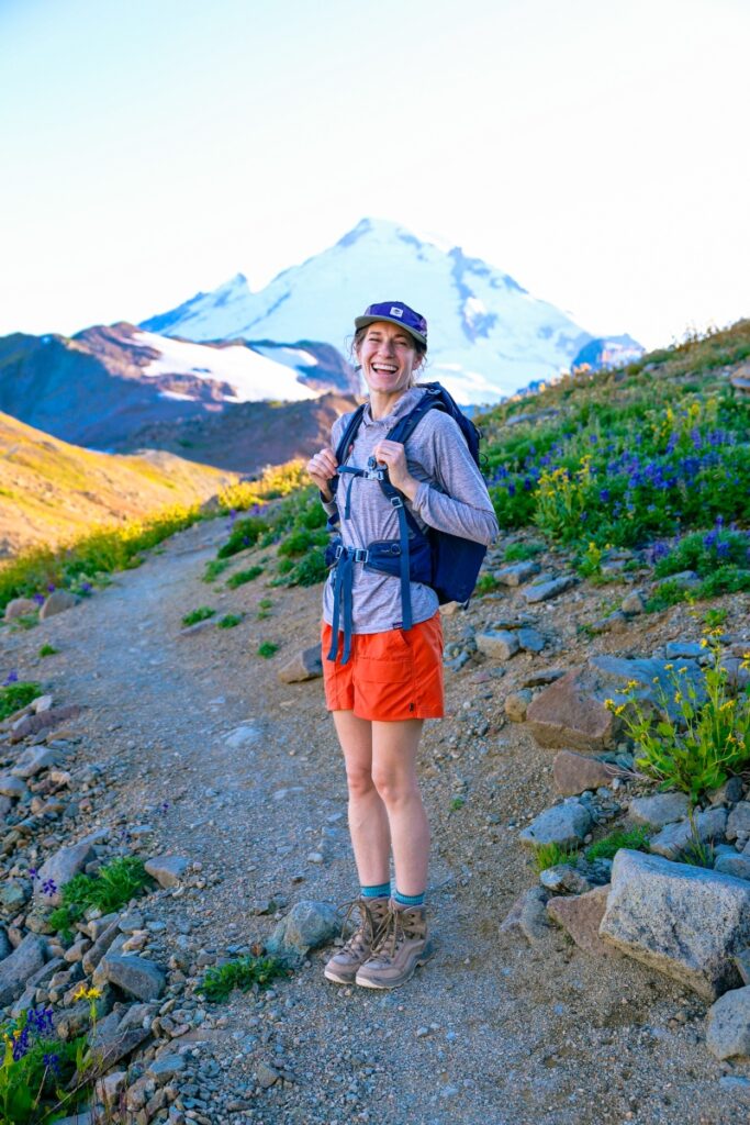 A women stands on a hiking trail wearing a backpack looking towards the camera with a mountain peak in the background