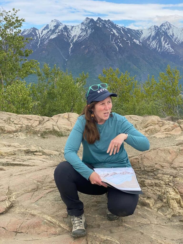 A woman is crouched down on a rock outcropping at the top of a mountain, holding a map and sharing information with her hiking mates out of frame