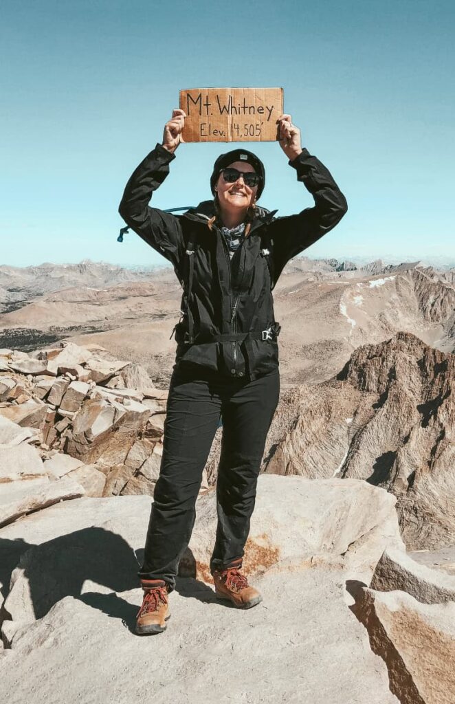 A hiker woman stands at the top of Mount Whitney with a cardoard sign that that reads "Mount Whitney Elev 14505'"