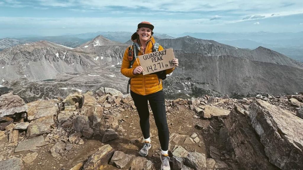 A hiker woman stands on a mountain with a sign that reads "Quandary Peak 147271' "