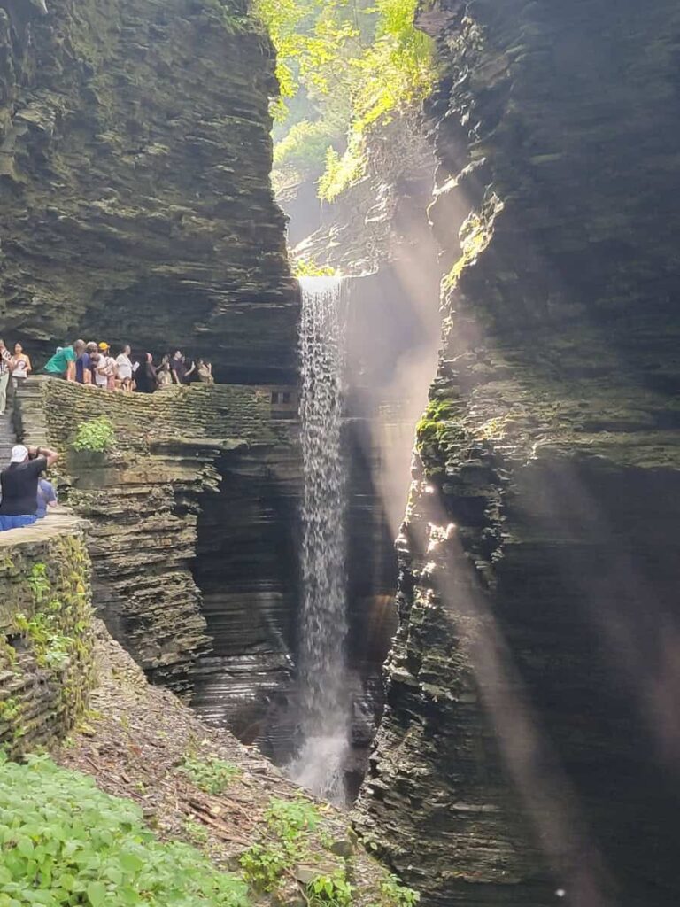 A tall waterfall flows over a walking path as visitors walk behind at Watkins Glen